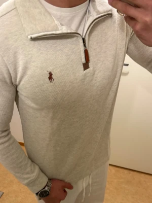 Beige stickad tröja med dragkedja - Snygg beige stickad tröja från Ralph Lauren med halv dragkedja och brunt läderdetalj vid zip. Klassisk broderad logga på bröstet. Långärmad modell i mjukt material, perfekt för lager på lager och chill stil.
