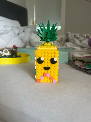 Lego ananas pennställ - Unikt pennställ byggt av Lego i form av en ananas. Kroppen är gul med ett glatt ansikte framtill och dekorerade sidor i pastellfärger som rosa, lila, blått och grönt. Toppen har gröna blad som ger en riktig ananasvibe. Perfekt för att förvara pennor eller småprylar på skrivbordet. De kommer isär och med massor av färgglada pluppar så man kan dekorera hur man vill