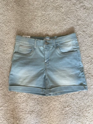 Ljusblå denimshorts - Säljer ett par ljusblå denimshorts med klassisk femficksdesign och uppvikta benslut. Shortsen har knapp och dragkedja framtill samt bälteshällor. Mjukt och skönt material.