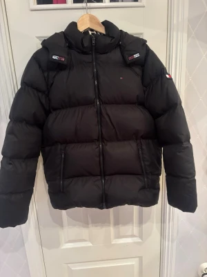 Svart pufferjacka Tommy Hilfiger - Snygg svart pufferjacka från Tommy Hilfiger med huva och dragkedja framtill. Jackan har två framfickor med dragkedja och diskreta logodetaljer på bröstet och ärmen. Perfekt för kalla dagar och riktigt skön att bära. Billigare vid snabb affär😊