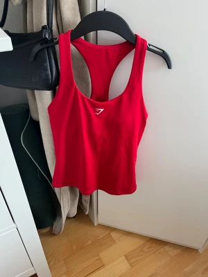 Oanvänt träningslinne från Gymshark - Snyggt rött träningslinne från Gymshark med brottarrygg och logga framtill. Tillverkat i ett stretchigt material av återvunnen polyester och elastan som andas bra under träningen. Perfekt för gymmet eller löprundan. Aldrig använt. Nypris 399 kr.
