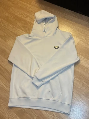 Prada tröja - Säljer en clean vit hoodie från Prada med den ikoniska trekantiga loggan på bröstet. Hoodien har huva med dragsko och ribbade muddar vid ärmar och nederkant. Perfekt för dig som gillar stilrena och exklusiva plagg.
