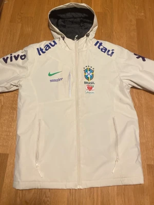 Vit Brasil Nike vindjacka med huva - Snygg vit vindjacka från Nike med Brasiliens landslagslogga och sponsormärken som Itaú och Vivo. Jackan har huva, hel dragkedja framtill och två sidofickor med dragkedja. Perfekt för dig som gillar fotboll och streetwear.