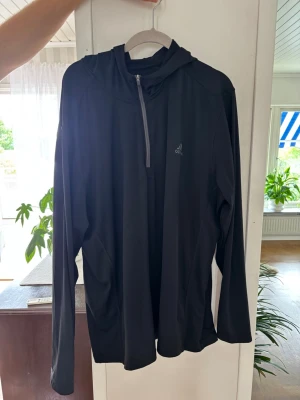 Svart hoodie från Adidas - Säljer en svart långärmad hoodie från Adidas med halv dragkedja framtill och diskret logga på bröstet. Tillverkad i ett lätt och stretchigt funktionsmaterial som passar perfekt till träning eller chill. Huva för extra komfort och stil.