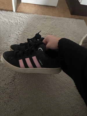 Svarta Adidas Campus sneakers - Säljer ett par svarta Adidas Campus sneakers med klassiska rosa ränder på sidorna och vit sula. Skorna har lågt skaft och snörning framtill. Perfekta för dig som gillar en stilren och sportig look.