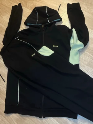 Boss dress - Säljer en svart hoodie från BOSS med ljusgröna och vita detaljer på ärmar och huva. Hoodien har dragkedja framtill, huva med snören och BOSS-logga på bröstet. Perfekt för dig som gillar stilrena och moderna plagg.