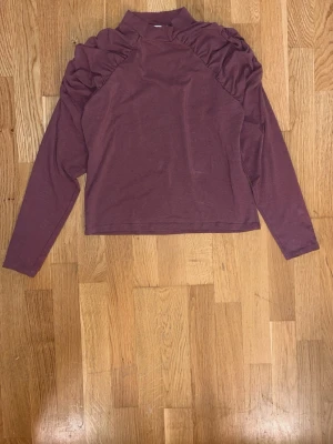 Plommonlila topp med puffdetaljer - Trendig långärmad topp i plommonlila med högt halsringning och snygga puffdetaljer längs axlarna. Tillverkad i ett mjukt, stretchigt material som sitter bekvämt. Perfekt för dig som vill ha en unik och stilren look.