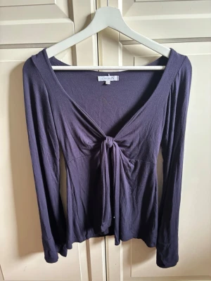 Mörklila långärmad topp med knyt - Säljer en mörklila långärmad topp från Design by Si. Toppen har v-ringning och snygg knytning framtill som ger en feminin touch. Tillverkad i mjukt material och passar perfekt till jeans eller kjol.