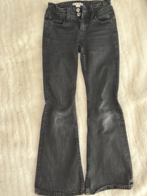 Svarta bootcut jeans med slitningar - Säljer ett par svarta bootcut jeans med snygga slitningar och två knappar i midjan. Jeansen har klassiska fem fickor och lätt utsvängda ben. Perfekta för dig som gillar en retro och avslappnad stil.