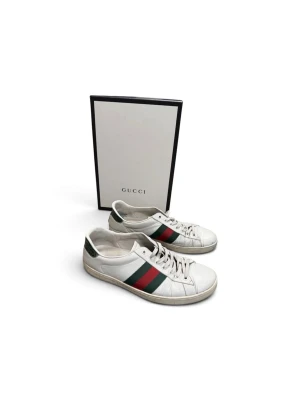 Gucci vita sneakers med ränder - Säljer ett par klassiska vita sneakers från Gucci med de ikoniska gröna och röda sidoremsorna. Skorna har lågt skaft, vita snören och detaljer i grönt på hälen. Tillverkade i läder och har en stilren, tidlös design som passar till det mesta.