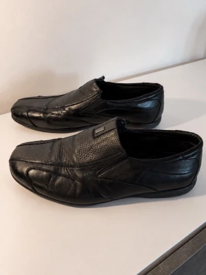 Svarta loafers i skinn från Bugatti - Snygga svarta loafers från Bugatti i äkta skinn. Skorna har en stilren design med diskreta sömmar och perforerat mönster på ovandelen. Låg sula i gummi med Bugatti-logga undertill. Perfekta för dig som gillar klassisk men modern stil.