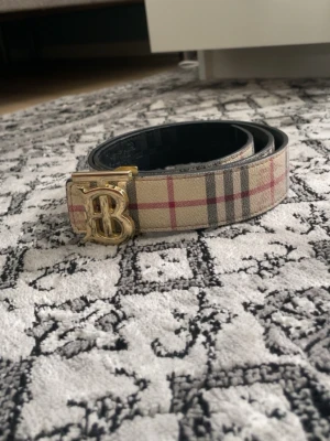 Beige rutigt bälte med guldspänne - Snyggt bälte i beige med klassiskt rutigt mönster och ett stort guldigt B-spänne framtill. Bältet har svarta insidor och passar perfekt för att lyfta vilken outfit som helst. Tillverkat i skinn för en exklusiv känsla.