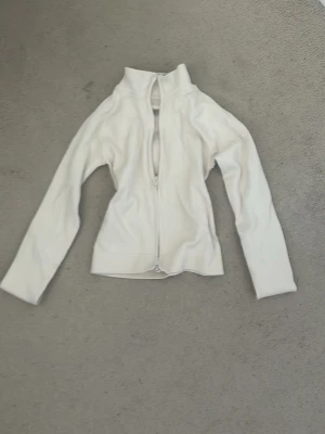 Vit ribbad träningströja - Säljer en vit ribbad tröja med dragkedja framtill och hög krage. Tröjan har långa ärmar och är tillverkad i ett stretchigt material som sitter snyggt på kroppen. Perfekt för en tränings outfit och att styla som lager på lager och enkel att styla.