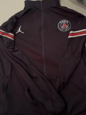 PSG x Jordan svart träningsjacka - Snygg svart träningsjacka från PSG x Jordan med hel dragkedja, vita och röda ränder på ärmarna samt PSG-logga och Jumpman-logga på bröstet. Tillverkad i ett mjukt och sportigt material som passar perfekt till träning eller chill.