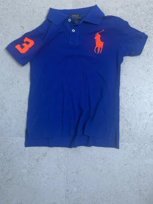 Blå pikétröja från Ralph Lauren för barn - Snygg blå pikétröja från Ralph Lauren med orange logga på bröstet och en stor orange siffra 3 på ärmen. Klassisk krage och knäppning med två knappar. Perfekt till en avslappnad men ändå stilren look. Storlek för sjuåringar.