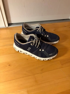 Marinblå On sneakers med vit sula - Snygga marinblå sneakers från On med vit, chunky sula och beige detaljer. Ovandelen är i mesh och syntetmaterial för bra andningsförmåga. Skorna har vita snören och låg profil som ger en sportig look. Perfekta för dig som gillar stilrena och bekväma sneakers.