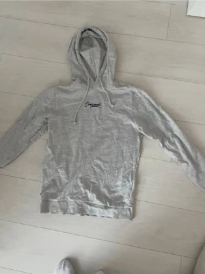 Grå hoodie från Jack and Jones  - Säljer en ljusgrå hoodie med huva och dragsnören. På bröstet finns en liten broderad text med ordet 'Original'. Hoodien har långa ärmar och en klassisk passform. Skriv vid frågor!  