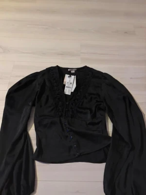 Svart blus med spetsdetaljer - Snygg svart blus i glansigt material med spetsdetaljer längs v-ringningen och knappar framtill. Blusen har långa, puffiga ärmar och en figurnära passform som ger en trendig look. Perfekt att styla med jeans eller kjol för en chic outfit.