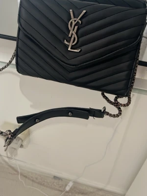 Svart YSL axelväska i läder - Snygg svart axelväska från YSL i quiltat läder med silverfärgad kedjerem och ikonisk YSL-logga framtill. Väskan har ett elegant, stilrent utseende och stängs med lock. Perfekt för dig som vill ha en trendig och klassisk accessoar.
