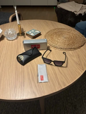 Svarta Ray-Ban solglasögon - Snygga svarta solglasögon från Ray-Ban med klassisk rektangulär form och mörka glas. Bågarna är i plast och kommer med originalfodral och putsduk. Perfekta för att höja din stil på soliga dagar.