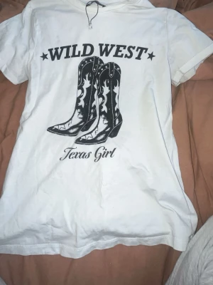 Vit Wild West t-shirt HB Sister - Vit t-shirt från HB Sister med tryck av cowboy boots och texten 'Wild West Texas Girl' på framsidan. Klassisk rund hals och korta ärmar. Regular fit-modell, perfekt för dig som gillar westernstil och statement-prints.