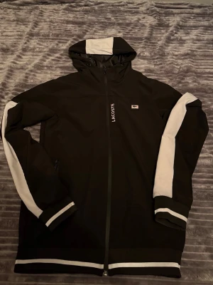 Svart jacka med vita detaljer Lacoste - Snygg svart hoodie från Lacoste med vita ränder på ärmarna, huvan och muddar. Dragkedja framtill och Lacoste-logga på bröstet. Perfekt för en sportig och avslappnad stil. Tillverkad i mjukt material för extra komfort.