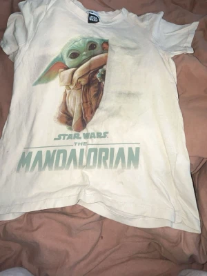 Star Wars Mandalorian t-shirt XS - Vit t-shirt från FB Sister med stort Mandalorian-tryck och Baby Yoda på framsidan. Klassisk rund hals och korta ärmar. Perfekt för dig som älskar Star Wars och vill ha en snygg och bekväm t-shirt med coolt motiv.