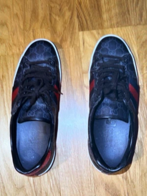 Bruna sneakers med röda och blå ränder - Snygga bruna sneakers med mönstrad ovandel och tydliga röda och blå ränder på sidorna. Skorna har vita sulor och svarta skosnören. Perfekta för dig som vill ha en stilren och trendig look till vardags.