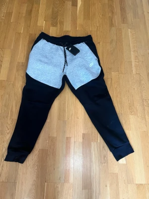 Nike mjukisbyxor svart och grå - Nike mjukisbyxor i svart och ljusgrått med dragsko i midjan och ribbade muddar vid bensluten. Byxorna har en modern, avsmalnande passform och Nike-logga på vänster sida. Perfekta för chill eller träning.