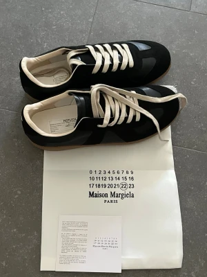 Strl.43 Svarta sneakers från maison margiela - Strl.43 Snygga svarta sneakers med beige gummisula och vita skosnören. Skorna har en klassisk låg profil och stilrena detaljer. Ovandelen verkar vara i syntet eller läder och sulan har ett mönster för bra grepp. Perfekta för en clean och tidlös look. Maison margielas