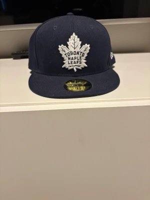Toronto Maple Leafs blå keps helt oanvänd - Snygg blå snapback-keps med Toronto Maple Leafs-logga broderad framtill och NHL-märke baktill. Köpt i officiella NHL butik i New York. se på sista bilden vilken storlek. Helt oanvänd