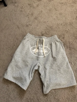 Gråa corteiz shorts  - Säljer ett par grå sweatshorts med vit snörning i midjan och tryckt vitt motiv framtill. Shortsen har avslappnad passform och är gjorda i mjukt bomullsmaterial, perfekta för chill dagar.