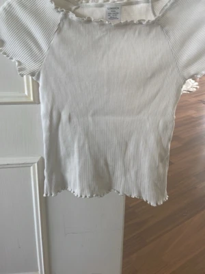 Vit ribbad topp från Lindex - Säljer en vit ribbad topp från Lindex med korta ärmar och vågig kant vid hals, ärmslut och nederkant. Toppen har ett diskret randigt mönster och är tillverkad i mjuk bomull, perfekt för en enkel och clean look.