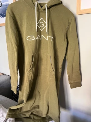 Olivgrön hoodieklänning från GANT - Säljer en olivgrön hoodieklänning från GANT med vit logga och grafiskt tryck på bröstet. Hoodien har huva med vita snören och en stor känguruficka framtill. Långa ärmar med ribbade muddar. Perfekt för en avslappnad och trendig look.