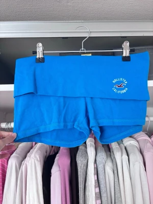 Blåa Hollister shorts med logga - Säljer ett par blåa shorts från Hollister med bred, vikbar linning och vit broderad logga framtill. Shortsen har en tight passform och är perfekta för varma dagar. Materialet känns mjukt och stretchigt, vilket gör dem riktigt bekväma.