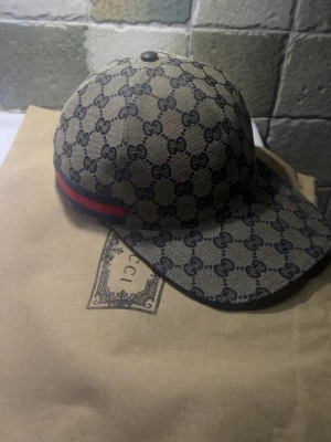 Gucci beige keps med GG-mönster - Säljer en beige keps från Gucci med klassiskt GG-monogram över hela kepsen och en röd-blå rand på sidan. Justerbar rem baktill i svart läder och guldfärgad spänne. Perfekt accessoar för dig som gillar exklusiv streetstyle.