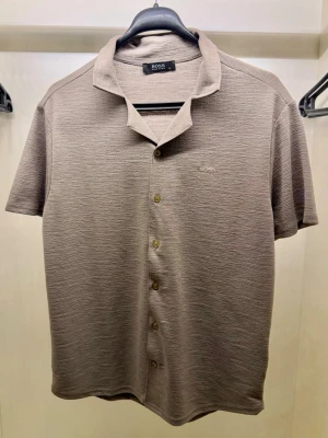 BOSS overshirt + shorts set - Säljer en stilren beige skjorta från BOSS med korta ärmar och klassisk krage. Skjortan har knappar framtill och diskret BOSS-logga broderad på bröstet. Materialet känns mjukt och har en lätt struktur som ger en modern look.