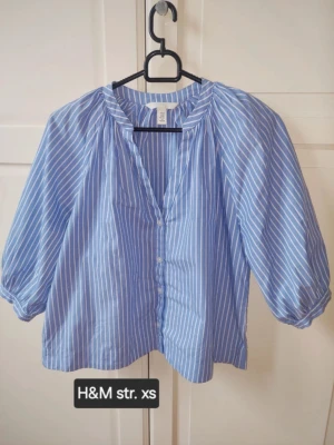 Blåvit randig blus från H&M - Snygg blå blus med vita vertikala ränder från H&M. Blusen har knappar framtill, lös passform och trekvartsärmar med lätt puff. Perfekt till jeans eller kjol för en fräsch look. Tillverkad i ett lätt och luftigt material.