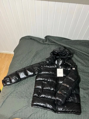 Muncler jacka - Säljer en svart, glansig dunjacka med huva och dragkedja framtill. Jackan har två sidofickor med dragkedja och breda, vadderade paneler som ger en puffig look. Perfekt för kalla vinterdagar och riktigt snygg till streetwear-stilen.