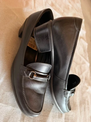 Svarta loafers med låg klack - Säljer ett par klassiska svarta loafers i skinn med låg klack och dekorativ metallspänne framtill. Skorna har rundad tå och diskreta sömmar längs kanterna för en stilren look. 