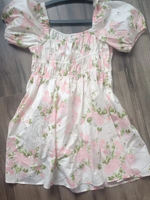 Blommig kortklänning med puffärm - Söt kortklänning med puffärmar och blommigt mönster i rosa och grönt på vit botten. Klänningen har en smockad överdel och fyrkantig ringning, vilket ger en romantisk och trendig look. Perfekt för varma dagar och somriga tillfällen.