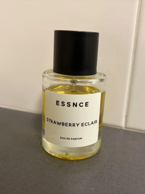 ESSNCE Strawberry Eclair parfym - En parfym från ESSNCE med doften Strawberry Eclair. Flaskan är cylindrisk i genomskinligt glas med svart lock och vit etikett med svart text. Innehållet är guldfärgat och flaskan har en stilren, modern design.