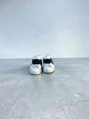 Vita Valentino sneakers med svart detalj - Snygga vita sneakers från Valentino med bred svart läderdetalj över sidan och klassisk snörning. Skorna har en låg profil och är tillverkade i läder för en exklusiv känsla. Perfekta för dig som vill ha en stilren och trendig look.