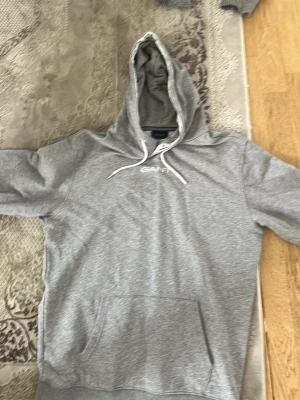 Grå hoodie från GANT med logga - Säljer en stilren grå hoodie från GANT med vit broderad logga på bröstet. Hoodien har huva med vita dragsnören och en stor magficka framtill. Tillverkad i mjukt sweatshirtmaterial som är perfekt för chill dagar.