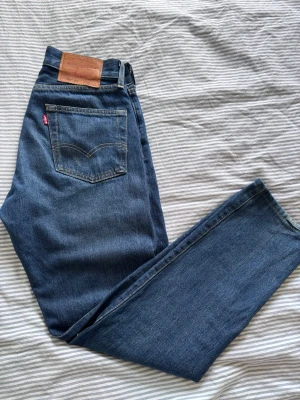 Levi's 501 blå raka jeans W28 L32 - Klassiska blå Levi's 501 jeans med rak passform och hög midja. Jeansen den ikoniska läderlappen bak i midjan. Tillverkade i slitstark denim med snygg tvätt och gul kontrastsöm.