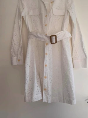 Vit broderad skjortklänning med bälte - Snygg vit skjortklänning i bomull med broderade detaljer och bälte i midjan. Klänningen har långa ärmar, knappar framtill och två bröstfickor. Perfekt för en fräsch och stilren look.