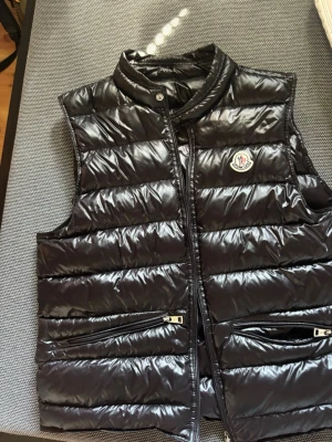 Svart dunväst från Moncler - Snygg svart dunväst från Moncler med glansig finish och klassisk logga på bröstet. Västen har ståkrage, dragkedja framtill och två fickor med dragkedja. Perfekt för lager på lager och ger en cool streetwear-look.