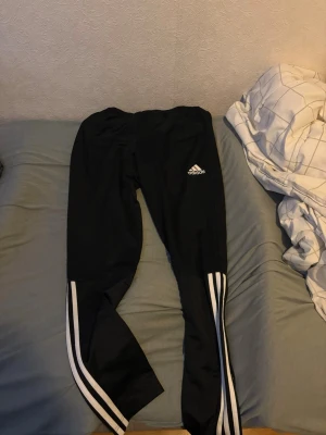 Svarta Adidas träningsbyxor - Säljer ett par svarta träningsbyxor från Adidas med klassiska vita ränder längs benen och Adidas-logga på låret. Byxorna har smal passform och är perfekta för gymmet eller löpning.