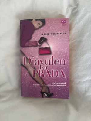 Djävulen bär Prada pocketbok - Säljer boken 'Djävulen bär Prada' av Lauren Weisberger. Pocketutgåva med rosa omslag och glittrig design, motiv av kvinna med handväska och högklackat. Perfekt för dig som gillar mode och underhållande skönlitteratur.