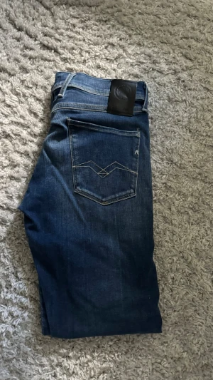 Blå slim fit jeans med bakficka - Säljer ett par blå slim fit jeans med klassisk femficksdesign och snygga detaljer på bakfickan. Jeansen har en mörkare tvätt med lätt slitning framtill och är tillverkade i denim. Perfekta för en avslappnad och trendig stil.